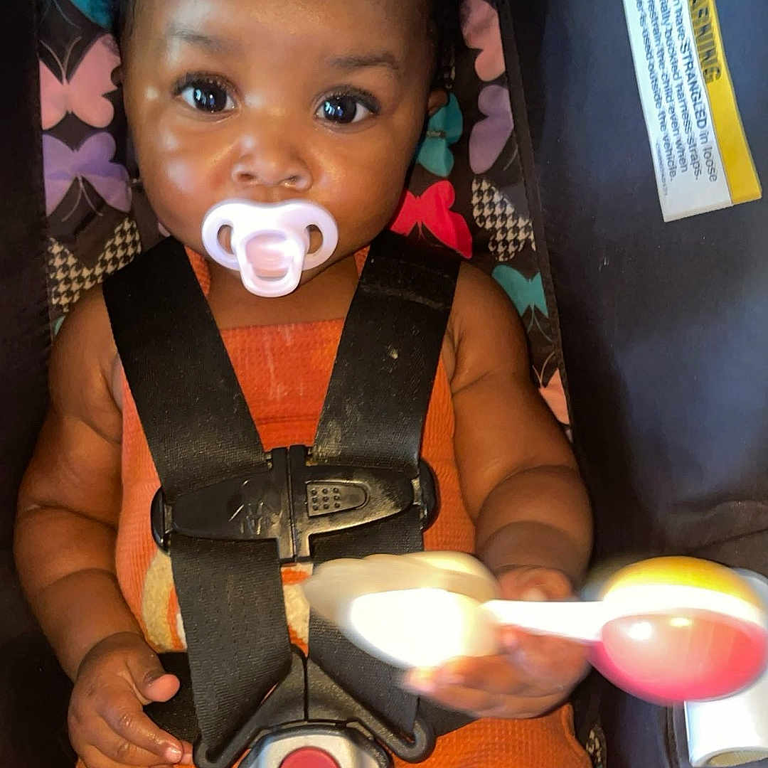 Milianah is registered to the contest to win money with this photo: baby, braided_hair, car_seat, child, closeup, colorful, cute, indoor, infant, orange_clothing, pacifier, patterned_background, playful, portrait, rattle_toy, safety_harness, seated, secure, smiling_eyes, young_child