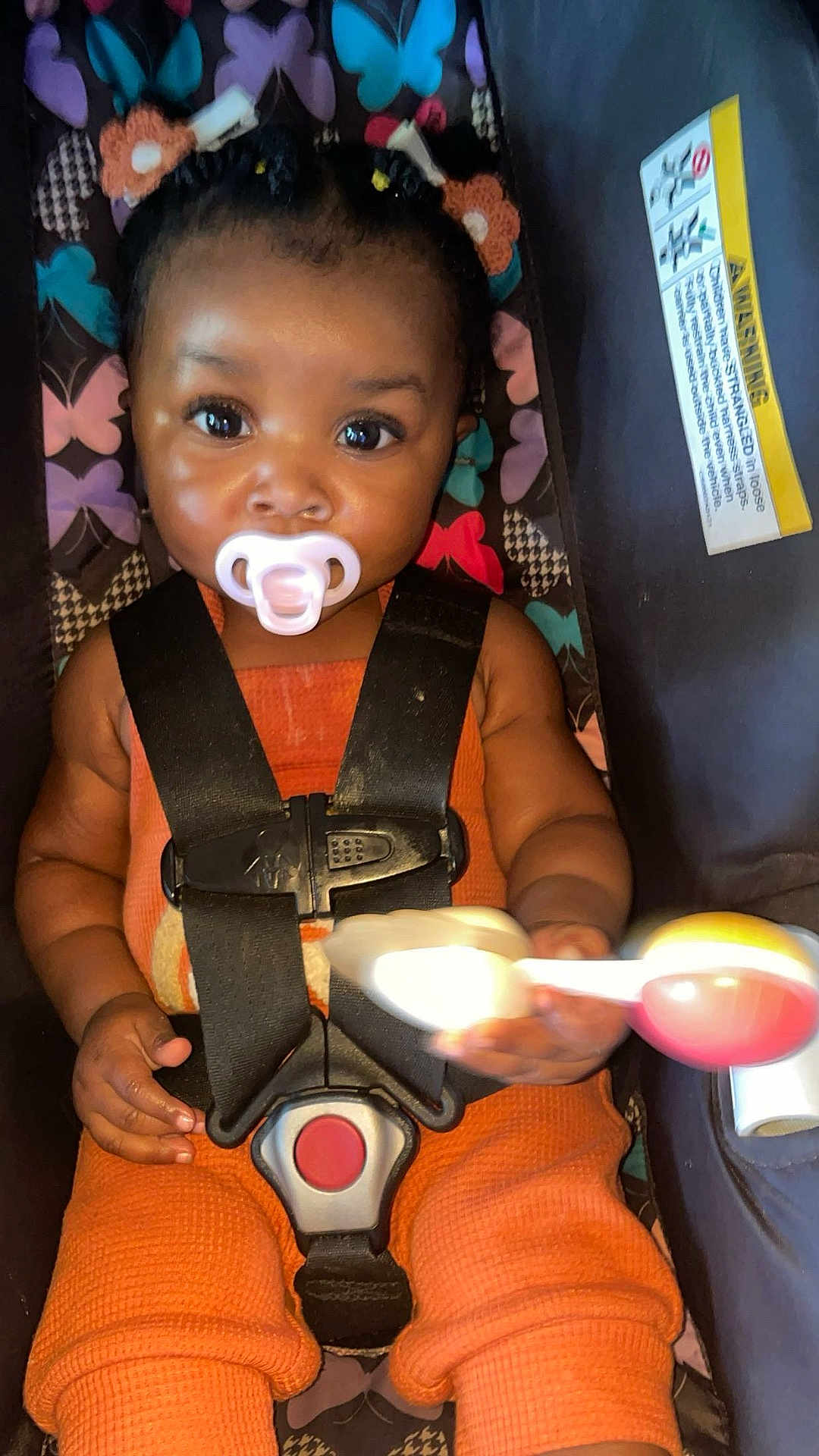 Milianah is registered to the contest to win money with this photo: baby, child, pacifier, car_seat, safety_harness, rattle_toy, orange_clothing, braided_hair, patterned_background, indoor, cute, infant, playful, colorful, closeup, portrait, smiling_eyes, seated, young_child, secure