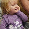 adult_arm, blonde_hair, casual_clothing, child, face, hand, happy, heart_pattern, high_chair, indoor, person, purple_shirt, sitting, smiling, table, tablecloth, tattoo, toddler, window, wood_paneling
