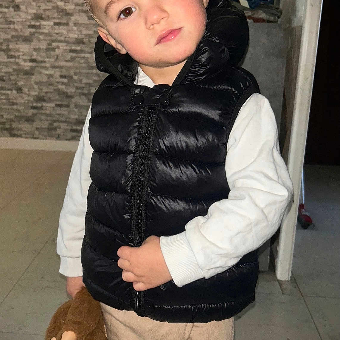 Maël participe au concours pour gagner de l'argent avec cette photo : beige_pants, black_jacket, brown_eyes, child, closup, cute, face, flash_photography, hand, indoor, patterned_wall, portrait, puffer_vest, standing, stuffed_monkey, tile_floor, toddler, toy, white_shirt, winter_clothing