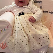 Aïtana a rejoint le concours — aidez-le/la à gagner de superbes lots ! baby, child, smile, happy, sitting, polka_dot, skirt, white_clothing, turban_hat, cushioned_chair, indoor, floor, wood_flooring, cute, portrait, infant, cozy, decor, text_on_chair, toddler