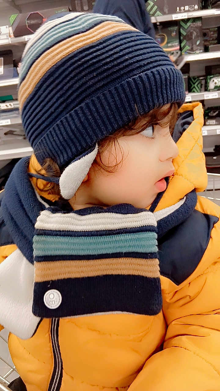 Awous a rejoint le concours — aidez-le/la à gagner de superbes lots ! toddler, child, hat, scarf, winter_clothing, yellow_jacket, curious, side_profile, indoor, shopping_cart, store, shelves, warm_clothing, striped, cute, face, person, fashion, bundle_up, cold_weather