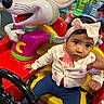 amusement_center, baby, blue_pants, bow_headband, cardigan, cartoon_mouse, child, colorful, headband, indoor, infant, kiddie_ride, playground, portrait, sitting, steering_wheel, sweater, toy, vehicle, wide_eyed