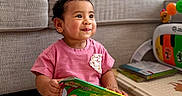 Ariana Castillo is registered to the contest to win money with this photo: activity_table, baby, book, bracelet, child, colorful_book, cozy, curly_hair, earring, furniture, indoors, pink_shirt, playroom, portrait, reading, sitting, smiling, sofa, toddler, toy