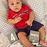 baby, infant, child, red_shirt, navy_shorts, pillow, blanket, blocks, month_block, number_block, indoor, cute, milestone, portrait, lying_down, soft_texture, checkered_pattern, curious_expression, hand, foot