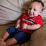 baby, child, smile, red_shirt, navy_shorts, couch, leather, indoor, happy, portrait, sitting, cute, infant, person, young_child, curly_hair, casual_clothing, relaxing, comfortable, home