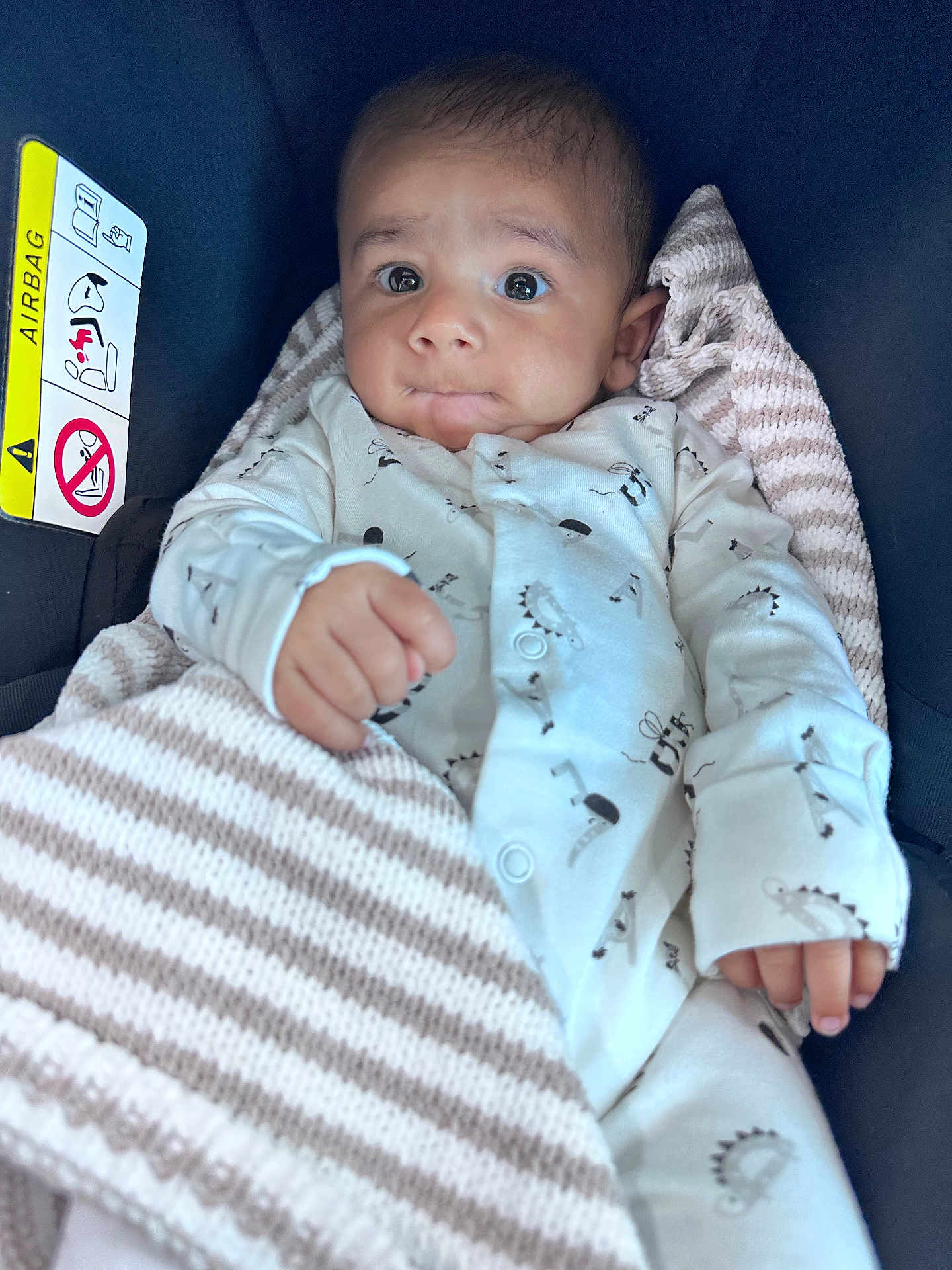 Gavin is registered to the contest to win money with this photo: baby, infant, car_seat, blanket, onesie, dinosaur_pattern, striped_blanket, face, wide_eyes, curious, hand, clothing, seatbelt_warning, indoor, portrait, child, cute, soft_texture, newborn, cozy