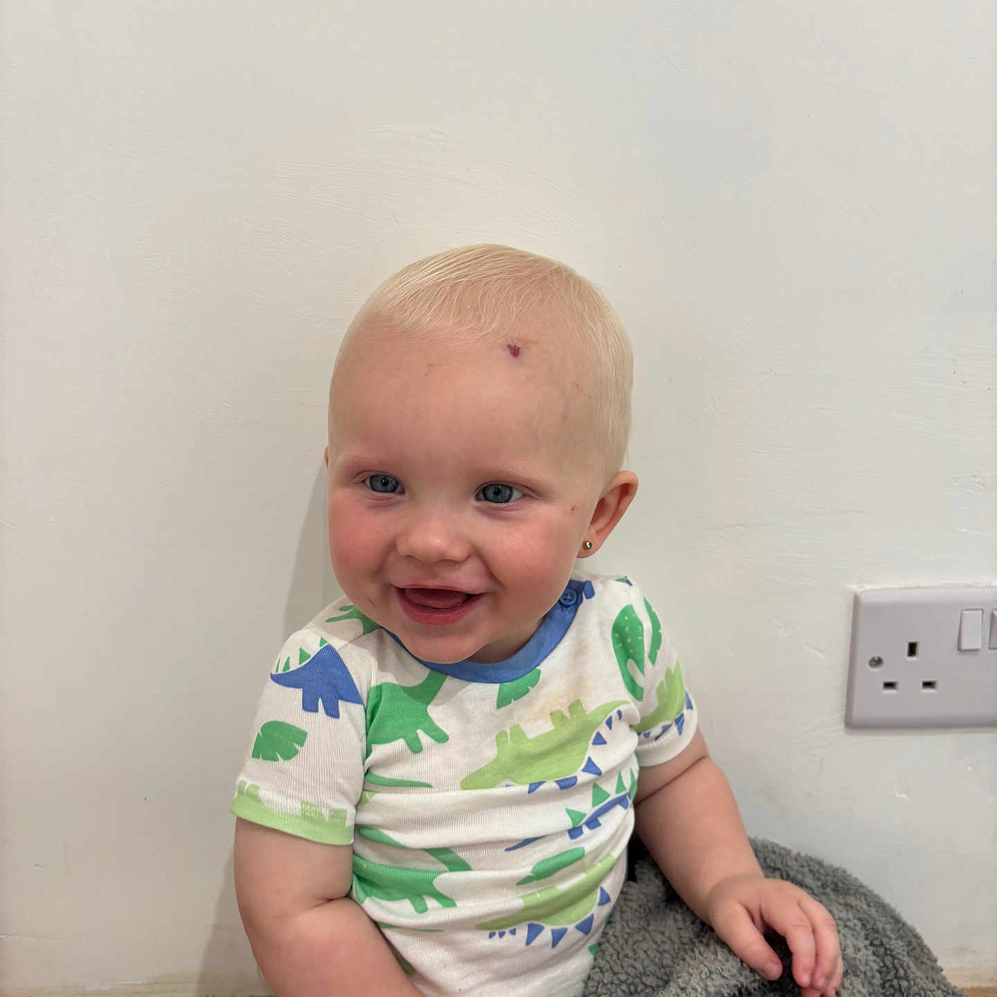Marley joined the competition — help win amazing prizes! baby, blanket, blue_eyes, casual_clothing, child, cute, dinosaur_pattern, earrings, happy, indoor, infant, playful, portrait, power_outlet, short_hair, sitting, smiling, toddler, white_wall, wooden_floor