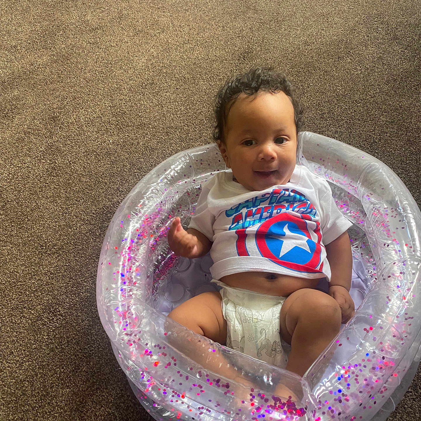 Mellow is registered to the contest to win money with this photo: baby, captain_america, carpet, child, curly_hair, cute, diaper, floor, glitter, indoor, infant, inflatable_pool, person, plastic, playful, relaxed, sitting, small_pool, smiling, tshirt