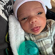Romello is registered to the contest to win money with this photo: newborn, baby, infant, child, portrait, face, eyes, tongue, pacifier, hat, winter_clothing, blanket, swaddled, cozy, close_up, indoors, seated, baby_carrier, textile, family_photo