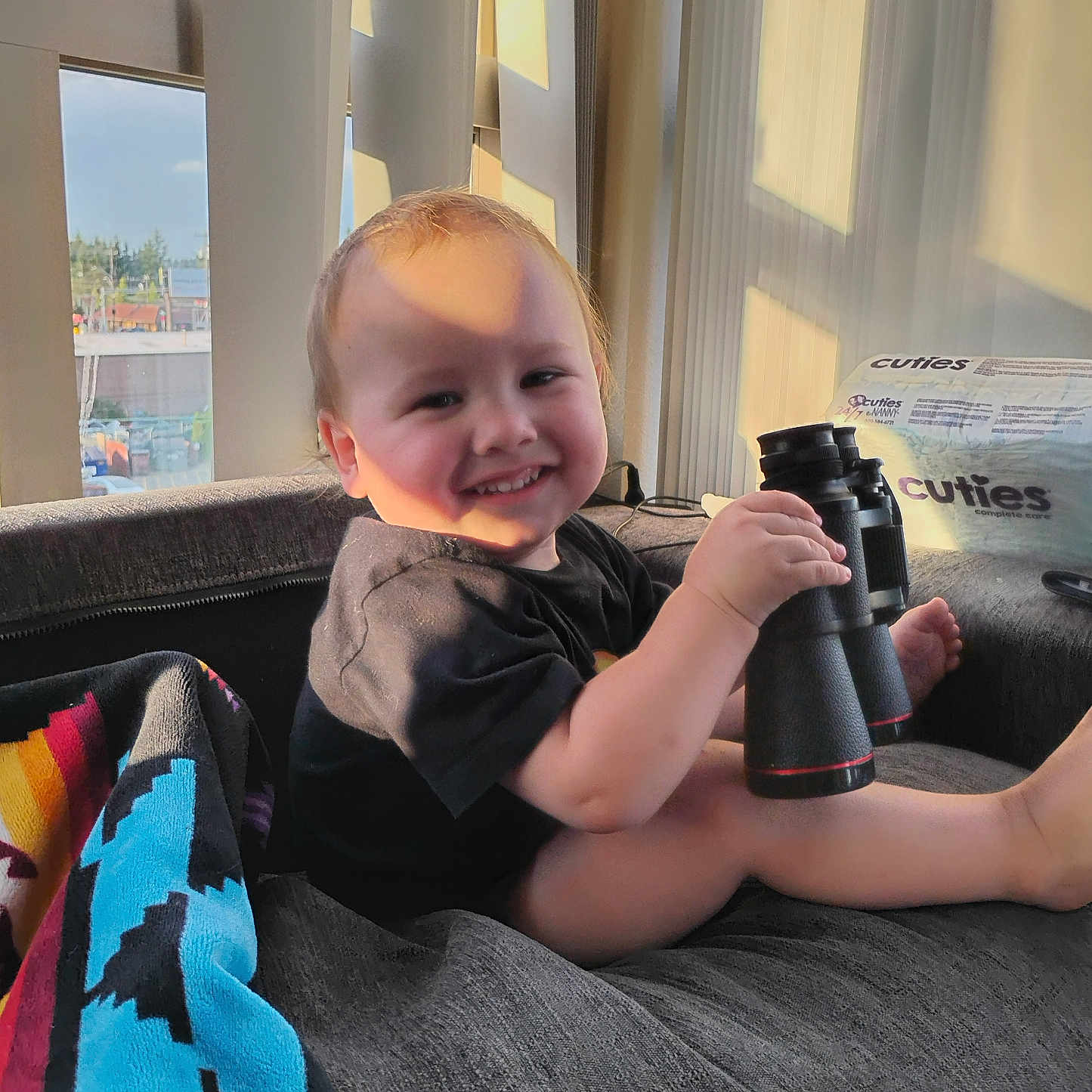 Ezekiel is registered to the contest to win money with this photo: binoculars, blanket, child, clothing, couch, curtains, face, furniture, happy, home, indoor, legs, person, relaxing, shadow, smile, sunlight, toddler, toy, window