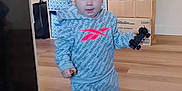 Ezekiel is registered to the contest to win money with this photo: toddler, child, indoor, toy_car, clothing, hoodie, pants, barefoot, flooring, storage_bins, box, water_bottles, wooden_floor, person, home, walking, face, blue_eyes, short_hair, casual