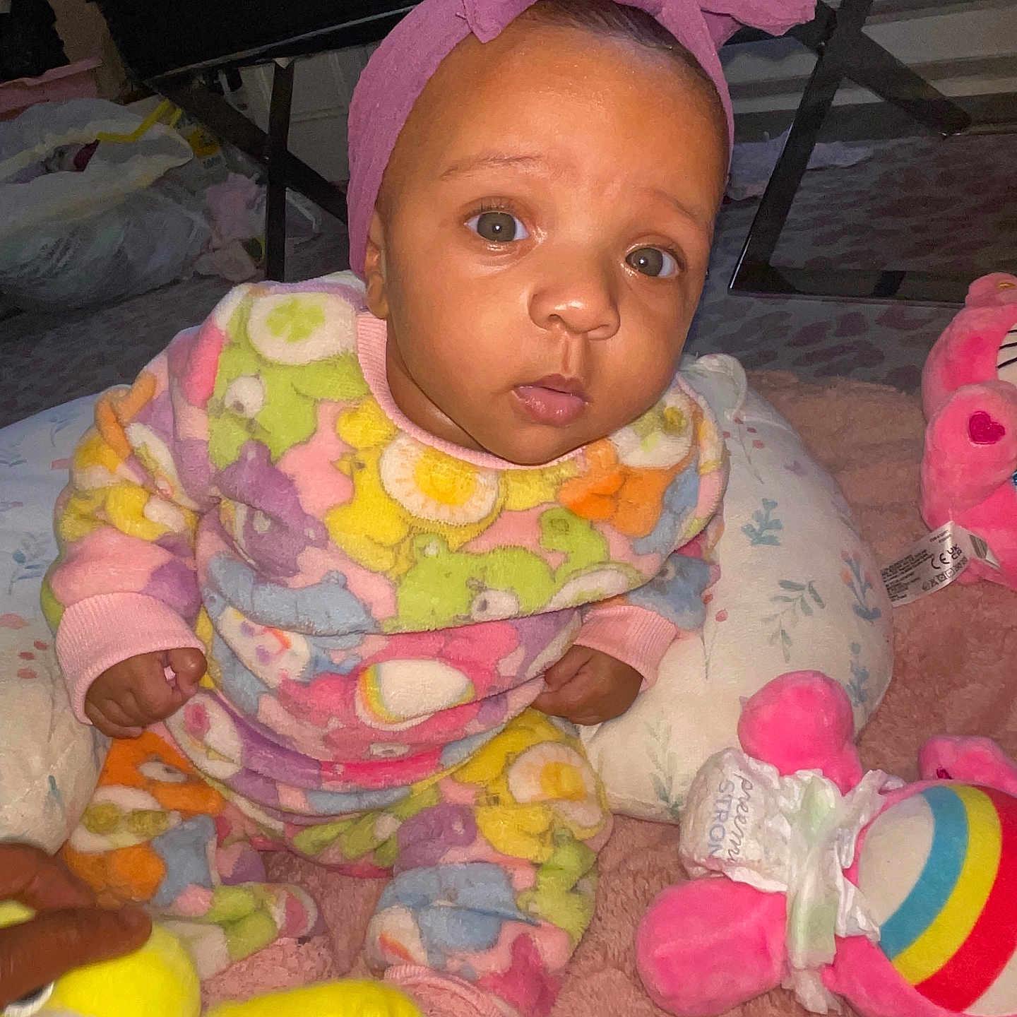 Kamora is registered to the contest to win money with this photo: baby, bright_colors, carpet, child, colorful_clothing, cozy, curious, cute, floor_seating, headwear, indoor, infant, patterned_outfit, pink_headband, playtime, plush_toys, portrait, sitting, soft_toys, young_child