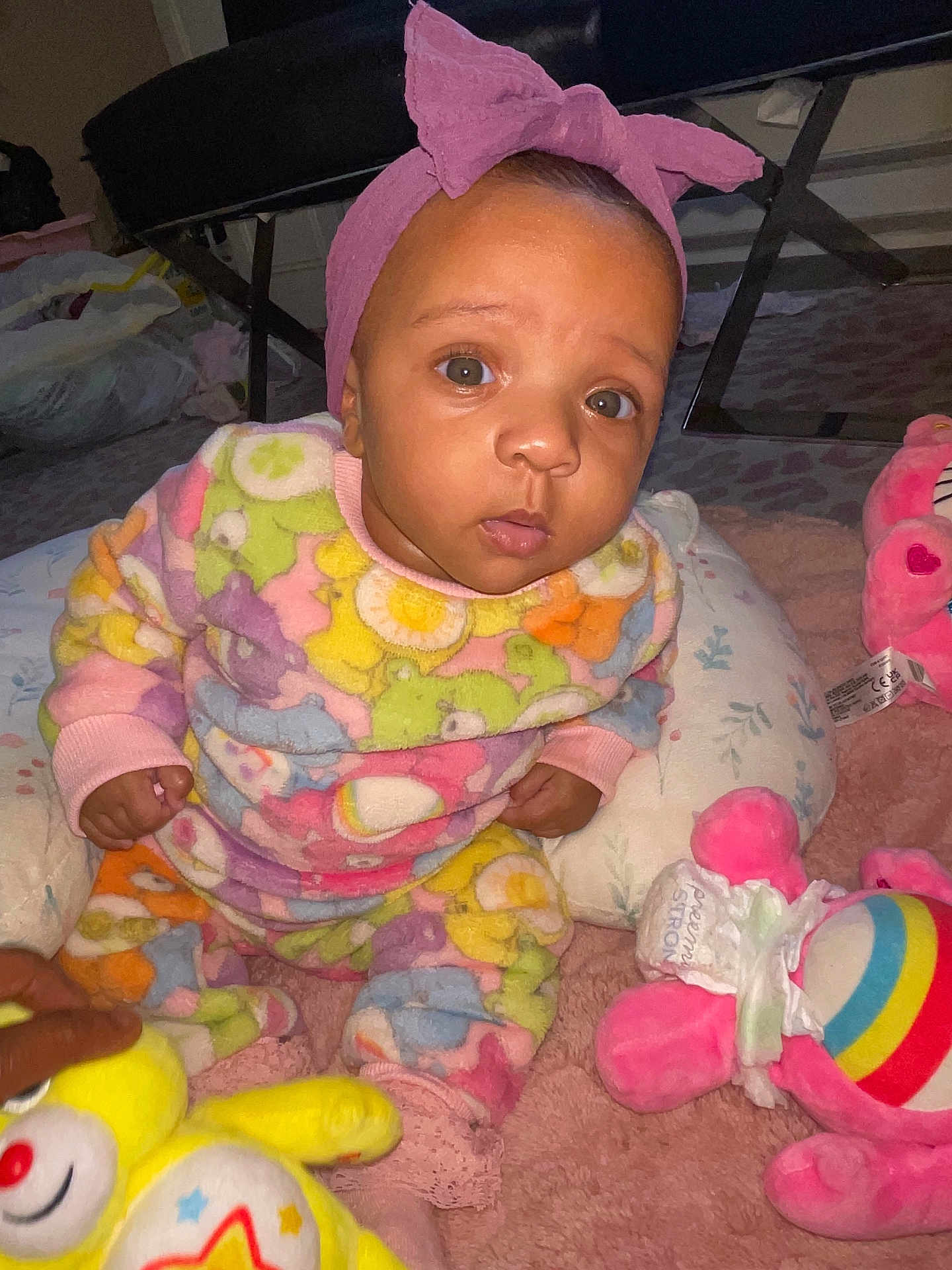 Kamora is registered to the contest to win money with this photo: baby, child, pink_headband, plush_toys, colorful_clothing, indoor, carpet, cute, sitting, curious, infant, soft_toys, patterned_outfit, headwear, young_child, playtime, floor_seating, bright_colors, cozy, portrait