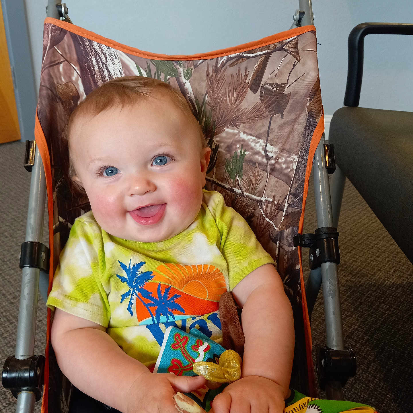 Charlie is registered to the contest to win money with this photo: baby, child, smiling, blue_eyes, stroller, camo_pattern, tie_dye_shirt, indoor, carpet, toy, blanket, happy, person, infant, clothing, seat, hand, face, cute, portrait