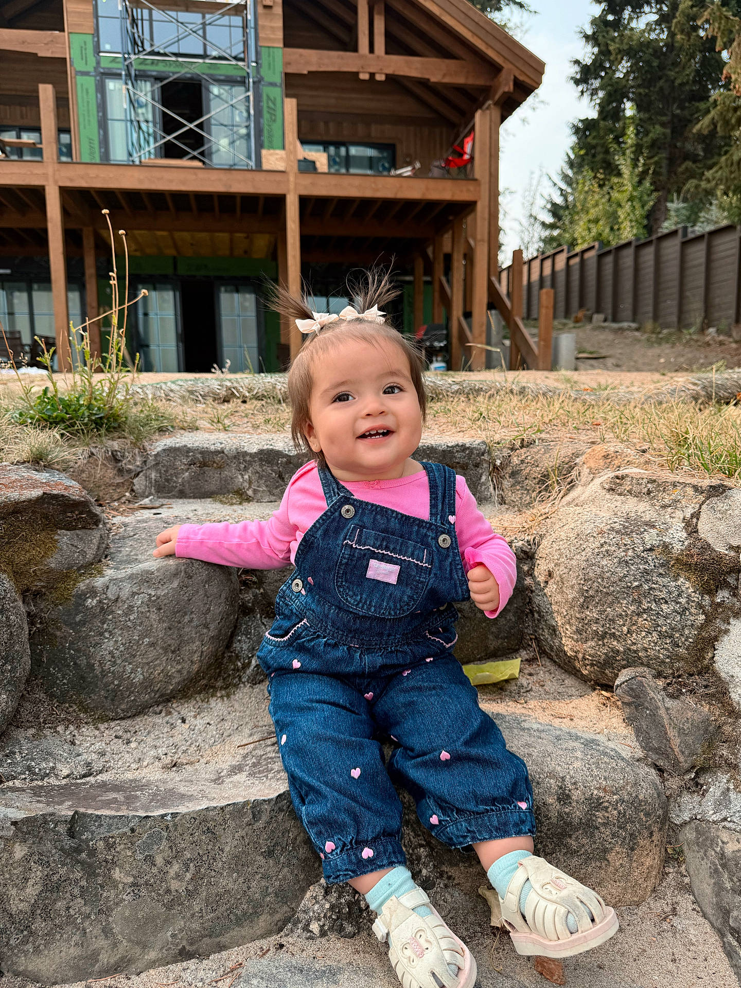 child, construction, cute, daylight, denim_overalls, grass, hair_bows, happy, nature, outdoor, person, pink_shirt, portrait, rocks, sandals, smiling, steps, toddler, trees, wooden_house