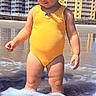 beach, beachfront, blue_sky, buildings, child, curly_hair, foam, ocean, outdoors, portrait, red_hair, seawater, standing, sunny, swimsuit, toddler, vacation, water, waves, yellow_clothing