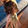 child, toddler, curly_hair, dress, floral_pattern, sunlight, portrait, indoor, wooden_floor, soft_light, side_profile, cute, young_child, person, curiosity, warm_colors, shadow, blurry_background, short_sleeves, standing