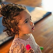 Sypha joined the competition — help win amazing prizes! child, toddler, curly_hair, indoor, toy, pink, dress, closeup, portrait, person, wooden_floor, blurred_background, cute, young_child, curious, holding, casual_clothing, face, small_toy, soft_lighting