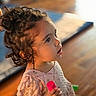child, toddler, curly_hair, indoor, toy, pink, dress, closeup, portrait, person, wooden_floor, blurred_background, cute, young_child, curious, holding, casual_clothing, face, small_toy, soft_lighting