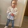 Erika is registered to the contest to win money with this photo: carpet, casual_clothing, child, curious_expression, cute, floral_pattern, footwear, girl, hair, hair_clips, hands_clasped, indoor, jeans, long_sleeve_shirt, person, portrait, standing, wall, white_sneakers, young