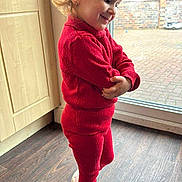 Erika is registered to the contest to win money with this photo: cabinet, casual, child, curly_hair, earrings, glass_door, happy, home, indoor, knitwear, natural_light, pose, red_clothing, smiling, sneakers, standing, toddler, topknot, window, wood_floor