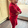 cabinet, casual, child, curly_hair, earrings, glass_door, happy, home, indoor, knitwear, natural_light, pose, red_clothing, smiling, sneakers, standing, toddler, topknot, window, wood_floor