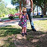 child, toddler, adult, outdoor, park, sunglasses, dress, floral_pattern, yellow_bow, smiling, standing, holding_hands, family, grass, trees, flowers, sidewalk, sunny, portrait, casual_clothing