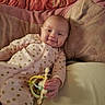 baby, infant, child, smiling, portrait, lying_down, pajamas, pink_clothing, polka_dots, toy, rattle, bed, pillows, blanket, bedding, indoor, home, cozy, happy, phone_screenshot