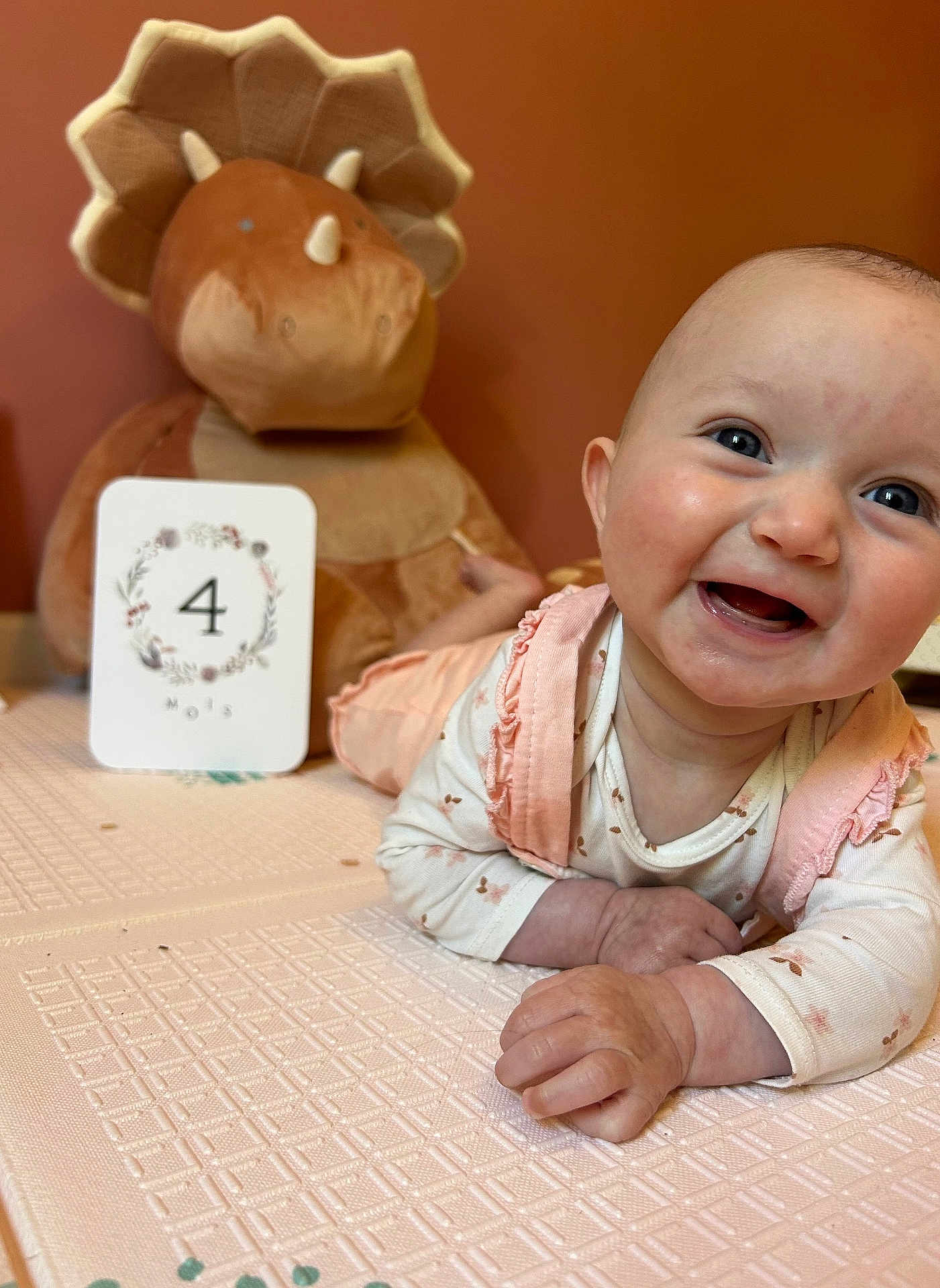 Anna participe au concours pour gagner de l'argent avec cette photo : baby, smile, infant, tummy_time, plush_toy, dinosaur, milestone_card, pink_clothing, white_clothing, happy, child, indoor, soft_toy, texture, face, person, cute, young_child, table, background
