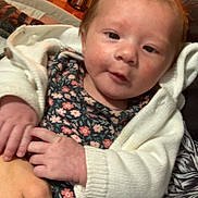 Sophia is registered to the contest to win money with this photo: baby, infant, child, person, hand, clothing, cardigan, floral_pattern, face, skin, indoor, furniture, wooden_floor, holding, portrait, young_child, expression, warm_lighting, home, cozy