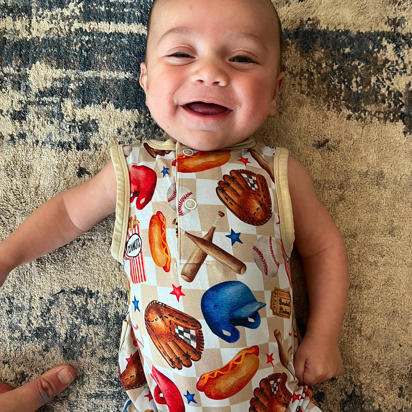 Elijah is registered to the contest to win money with this photo: apparel, baby, baseball, bat, carpet, cheerful, child, cute, glove, hand, happy, helmet, hot_dog, indoors, infant, lying_down, patterned_clothing, person, playful, smiling