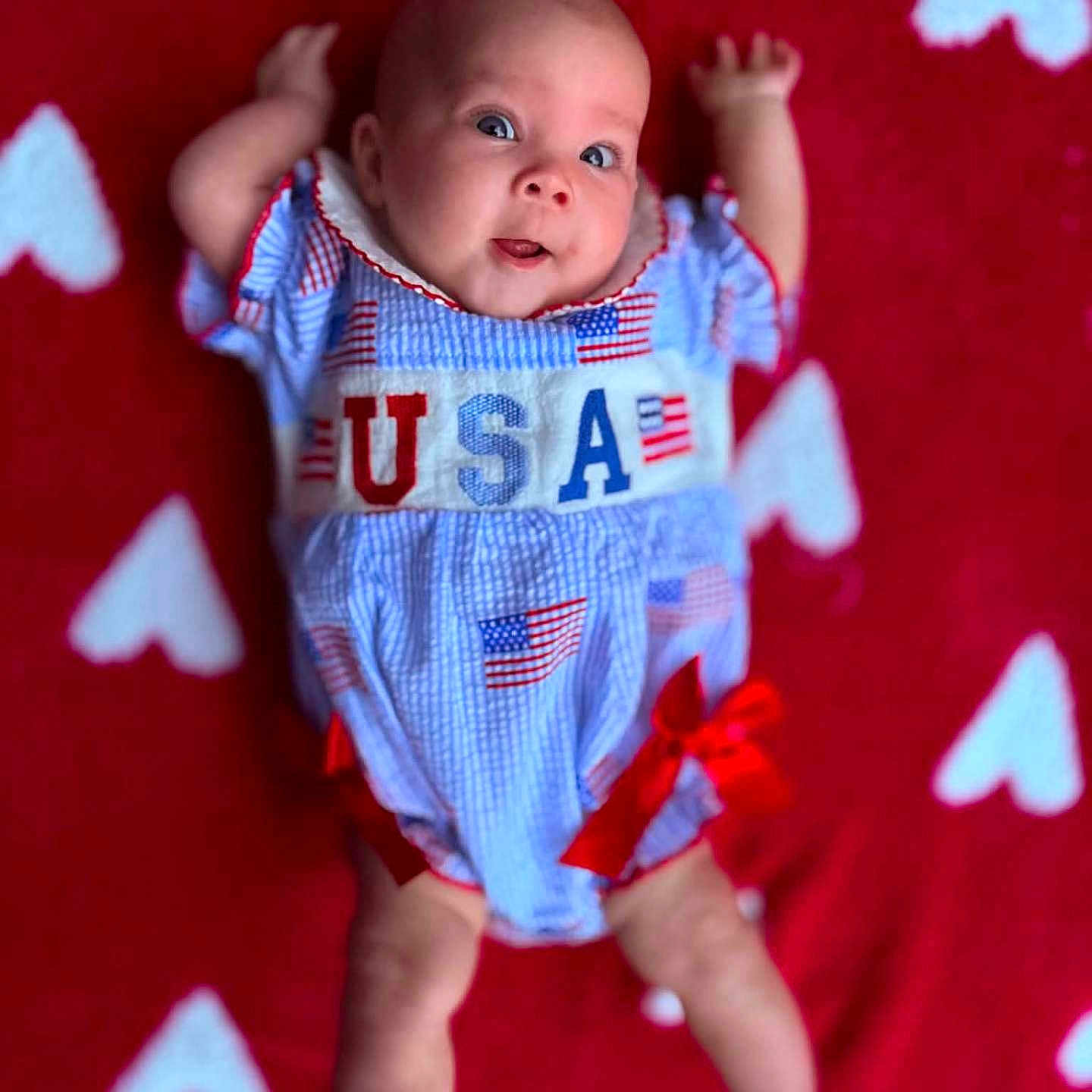 Violet joined the competition — help win amazing prizes! baby, infant, child, red_blanket, white_hearts, usa_outfit, flags, blue_clothing, red_bows, cute, smiling, lying_down, small_arms, small_legs, face, eyes, mouth, happy, indoor, portrait