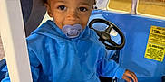 Jaylen is registered to the contest to win money with this photo: child, toddler, pacifier, blue_hoodie, curly_hair, play_car, steering_wheel, hand, window, door_frame, indoor, sign, martial_arts, kickboxing, jiu_jitsu, floor, toy, cute, portrait, smiling
