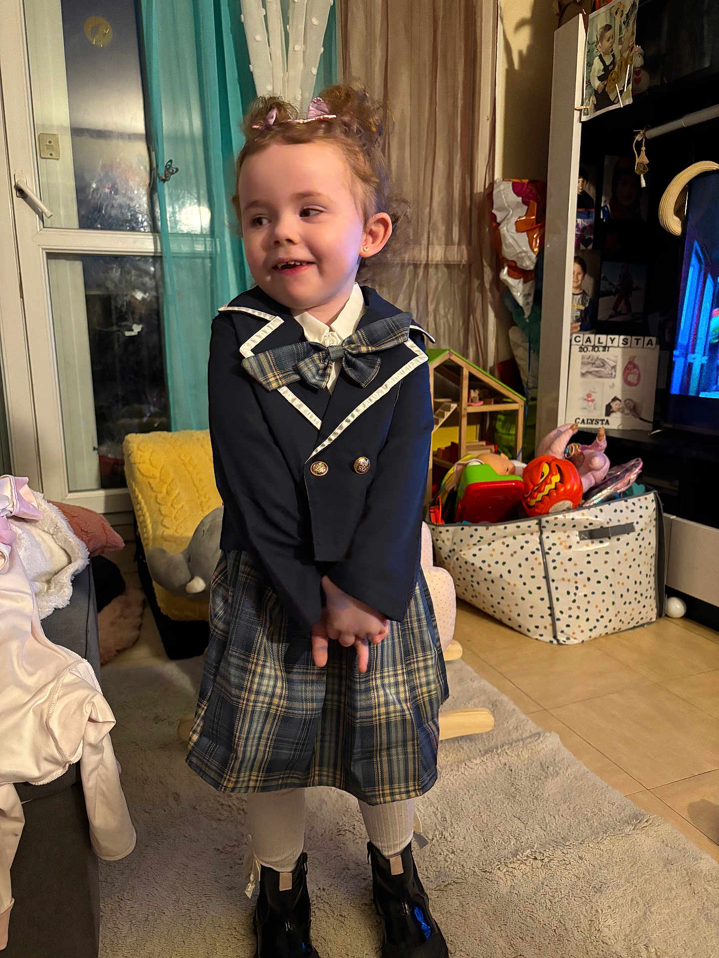 Calysta participe au concours pour gagner de l'argent avec cette photo : child, girl, plaid_skirt, blazer, bow, smile, indoor, carpet, toy_box, toys, cushion, window, curtains, boots, clothing, home, furniture, floor, person, portrait