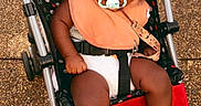 Royal is registered to the contest to win money with this photo: baby, pacifier, stroller, infant, child, outdoor, footwear, bib, polka_dot, mickey_mouse_pattern, curly_hair, black_hair, barefoot, seatbelt, fabric, ground, daylight, person, cute, weekend