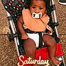 baby, pacifier, stroller, infant, child, outdoor, footwear, bib, polka_dot, mickey_mouse_pattern, curly_hair, black_hair, barefoot, seatbelt, fabric, ground, daylight, person, cute, weekend