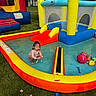 child, toddler, baby, outdoor, backyard, splash_pad, inflatable_pool, water, water_slide, bounce_house, playground, toys, ball, basketball, summer, grass, residential, portrait, sitting, colorful