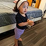 Rose joined the competition — help win amazing prizes! child, toddler, portrait, headband, standing, barefoot, holding_object, phone_case, bedroom, bed, wardrobe, wood_floor, indoor, curious, looking_sideways, black_shirt, purple_shorts, brown_hair, face, home