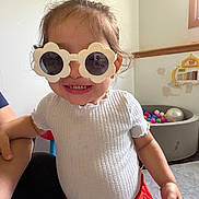 Rose is registered to the contest to win money with this photo: child, toddler, smiling, sunglasses, flower_glasses, portrait, indoor, playroom, toy, xylophone, ball_pit, colorful_balls, white_shirt, red_skirt, standing, adult_hand, furniture, window, wall, happy