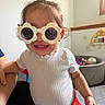 child, toddler, smiling, sunglasses, flower_glasses, portrait, indoor, playroom, toy, xylophone, ball_pit, colorful_balls, white_shirt, red_skirt, standing, adult_hand, furniture, window, wall, happy