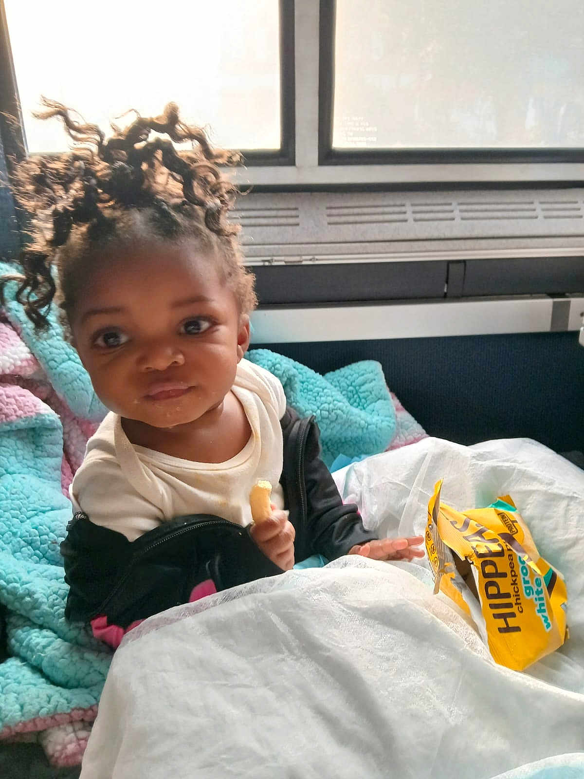 Yemaya joined the competition — help win amazing prizes! toddler, child, curly_hair, blanket, snack, food, indoor, window, seat, clothing, jacket, face, expression, person, young_child, cozy, curious, yellow_bag, white_cover, soft_texture