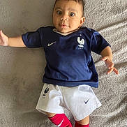 Kassim participe au concours pour gagner de l'argent avec cette photo : baby, child, soccer_uniform, blue_shirt, white_shorts, red_socks, lying_down, blanket, indoor, cute, sport, young_child, portrait, person, infant, athletic_wear, sporty, smiling, happy, face