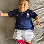 baby, child, soccer_uniform, blue_shirt, white_shorts, red_socks, lying_down, blanket, indoor, cute, sport, young_child, portrait, person, infant, athletic_wear, sporty, smiling, happy, face