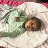 baby, child, blanket, bedspread, pink, white, green_sweater, gray_pants, lying_down, indoors, infant, person, face, head, curly_hair, cozy, resting, cute, young_child, casual_clothing