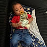 baby, child, pacifier, money, dollar_bills, red_shirt, blue_jeans, blanket, dinosaur_pattern, couch, footwear, black_shoes, indoor, person, infant, clothing, lying_down, holding, cute, casual