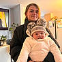 Christopher is registered to the contest to win money with this photo: baby, woman, indoor, hat, pom_pom, jacket, smile, face, person, bracelet, plant, refrigerator, television, candle, home, cozy, warm_clothing, holding, young_adult, dim_lighting