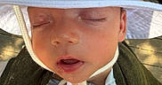 Hunter is registered to the contest to win money with this photo: baby, sleeping, hat, white_hat, chin_strap, sunlight, wooden_deck, closeup, infant, skin, peaceful, child, outdoor, green_cloth, carrier, face, portrait, hand, relaxing, toddler