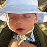 baby, sleeping, hat, white_hat, chin_strap, sunlight, wooden_deck, closeup, infant, skin, peaceful, child, outdoor, green_cloth, carrier, face, portrait, hand, relaxing, toddler