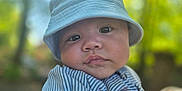 Theo is registered to the contest to win money with this photo: baby, child, face, hat, bucket_hat, striped_shirt, hand, outdoor, nature, greenery, portrait, closeup, infant, young_child, person, cute, skin, expression, holding, daylight